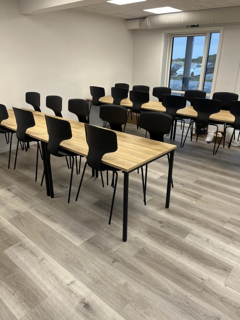 Kantine med lange borde i lyst træ, sorte stole og lyst gulv